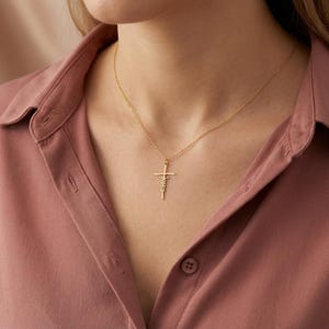 May include: A gold cross pendant necklace with the word "Faith" inscribed on the cross. The necklace is worn on a person wearing a pink button-down shirt.