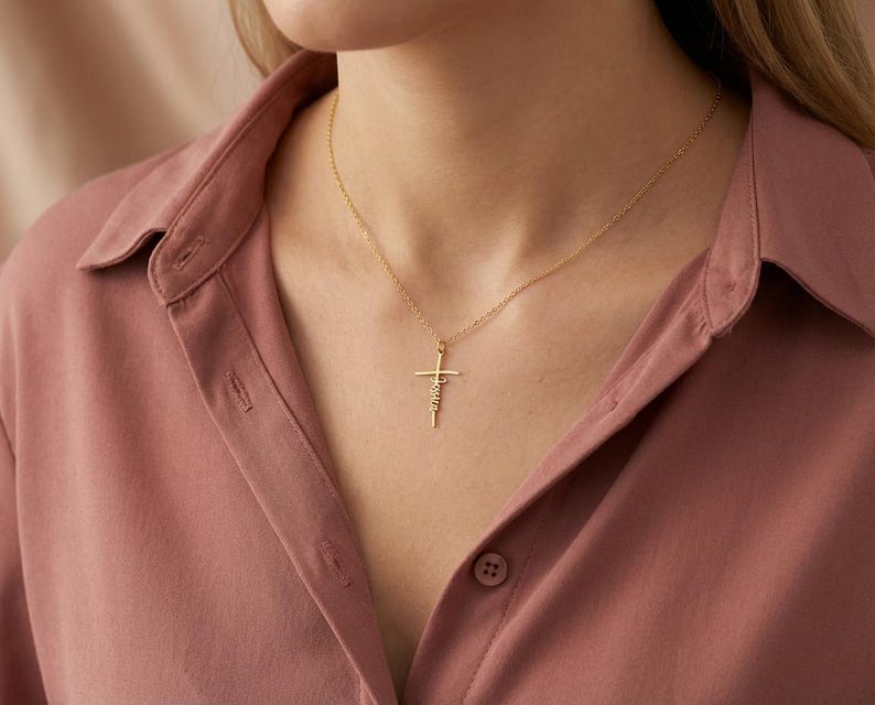 May include: A gold cross pendant necklace with the word "Faith" inscribed on the cross. The necklace is worn on a person wearing a pink button-down shirt.