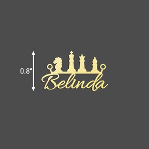 May include: Gold-coloured metal cutout of a chess piece crown with the name Belinda underneath. The crown is 0.8 inches tall.