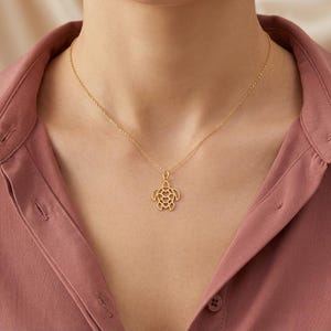 May include: A gold-colored necklace with a turtle pendant. The pendant is a cutout design, showcasing the turtle's shape. The necklace is worn against a pink shirt.