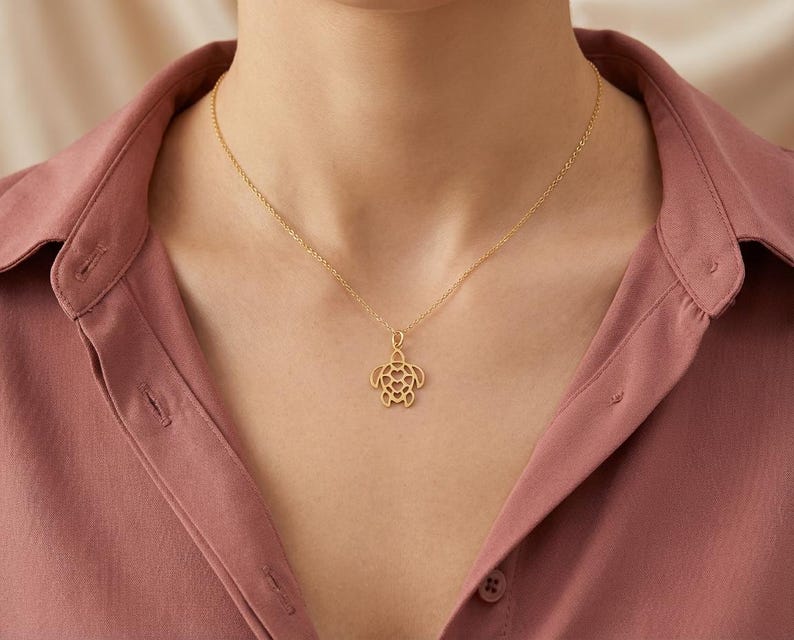 May include: A gold-colored necklace with a turtle pendant. The pendant is a cutout design, showcasing the turtle's shape. The necklace is worn against a pink shirt.