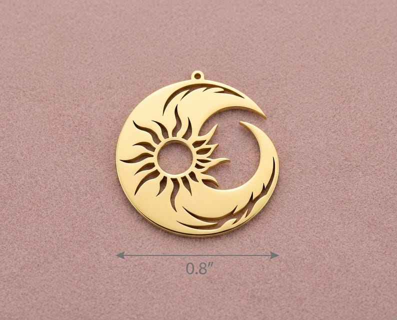 Op de afbeelding: Een goudkleurige hanger met een zon- en halve maan ontwerp. De zon staat in het midden, met stralen die naar buiten uitsteken. De maan buigt zich om de zon. De hanger heeft een diameter van 2,03 cm. Hij heeft een kleine lus aan de bovenkant om op te hangen.
