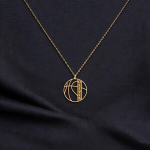 May include: A gold-colored necklace featuring a basketball design pendant. The pendant has the name "OLIVIA" vertically inscribed. The necklace is displayed on a dark blue fabric background.
