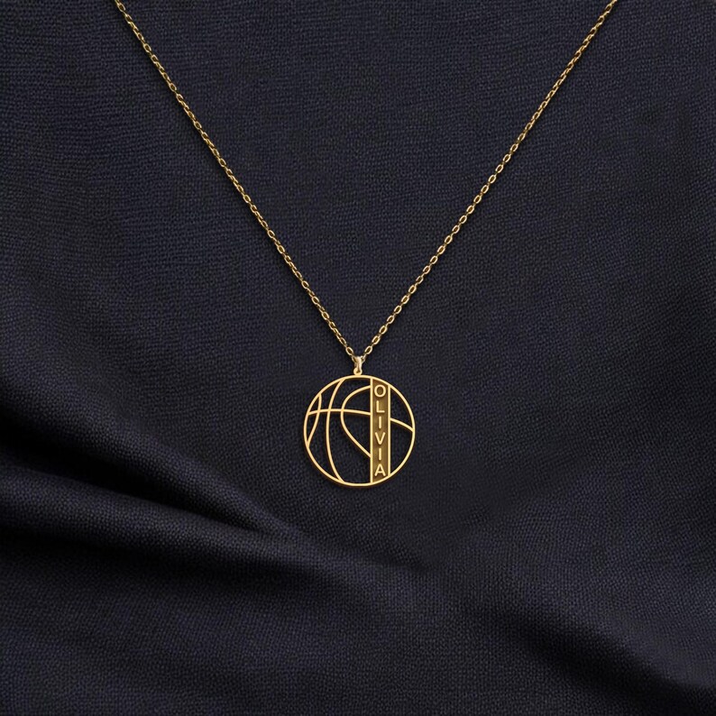 May include: A gold-colored necklace featuring a basketball design pendant. The pendant has the name "OLIVIA" vertically inscribed. The necklace is displayed on a dark blue fabric background.