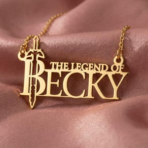 May include: Gold-colored necklace with the text "THE LEGEND OF BECKY" and a sword design. The necklace is displayed on a pink satin fabric. The pendant is a cutout design with a sword integrated into the letters.