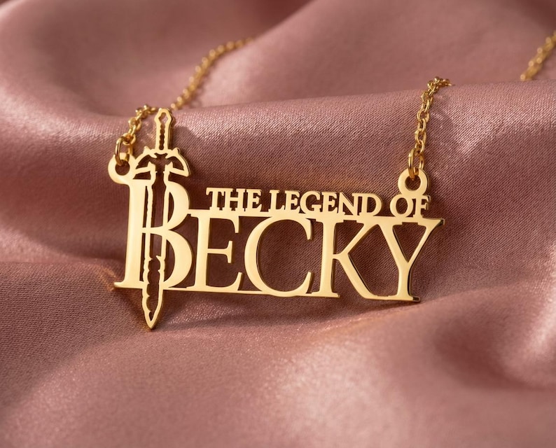 May include: Gold-colored necklace with the text "THE LEGEND OF BECKY" and a sword design. The necklace is displayed on a pink satin fabric. The pendant is a cutout design with a sword integrated into the letters.