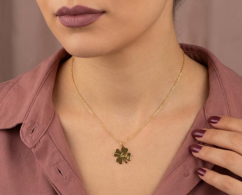 Pode incluir: Um colar dourado com um pingente de trevo de quatro folhas. O pingente tem a inscri&ccedil;&atilde;o "lucky". O colar &eacute; usado por uma pessoa vestindo uma camisa rosa.