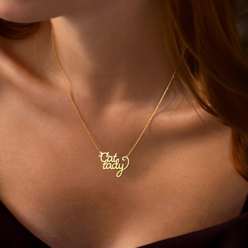 Peut inclure: Un collier en or avec un pendentif qui dit "Cat Lady" en &eacute;criture cursive. Le pendentif est en forme de t&ecirc;te de chat.