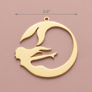 May include: A gold-colored pendant featuring a mermaid design. The pendant is approximately 0.8 inches in diameter and depicts a mermaid swimming within a circular frame. The mermaid has flowing hair and a detailed tail. The pendant has a loop at the top for hanging.