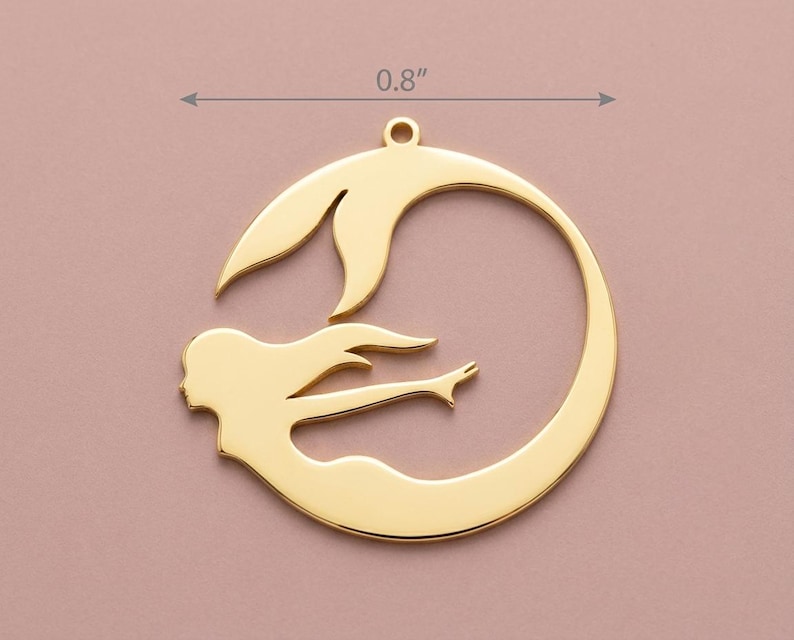 May include: A gold-colored pendant featuring a mermaid design. The pendant is approximately 0.8 inches in diameter and depicts a mermaid swimming within a circular frame. The mermaid has flowing hair and a detailed tail. The pendant has a loop at the top for hanging.