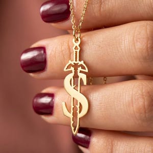 May include: A gold-colored pendant necklace featuring a sword design with a stylized letter "S" incorporated into the blade. The necklace is held by a person with burgundy nail polish. The chain is delicate and the pendant is detailed.