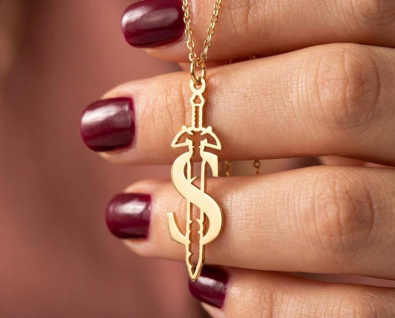May include: A gold-colored pendant necklace featuring a sword design with a stylized letter "S" incorporated into the blade. The necklace is held by a person with burgundy nail polish. The chain is delicate and the pendant is detailed.