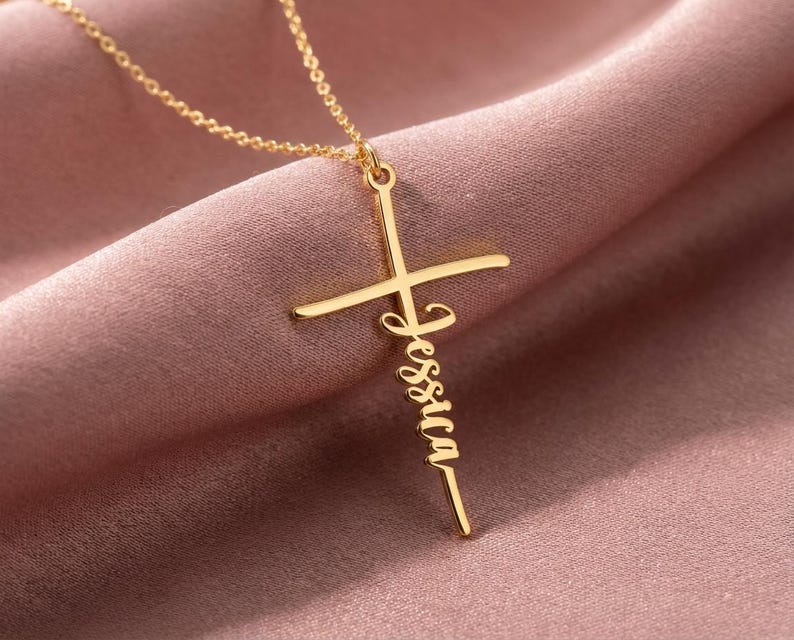 May include: A gold-coloured cross pendant necklace with the name "Jessica" inscribed vertically. The necklace is displayed on a pink fabric background. The cross has a classic design with a horizontal and vertical bar.