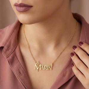 May include: A gold-colored necklace with the name "Marin" in block letters, with the words "THE LEGEND OF" above it. The necklace has a sword and shield design on the left side. The necklace is worn on a person wearing a pink shirt.