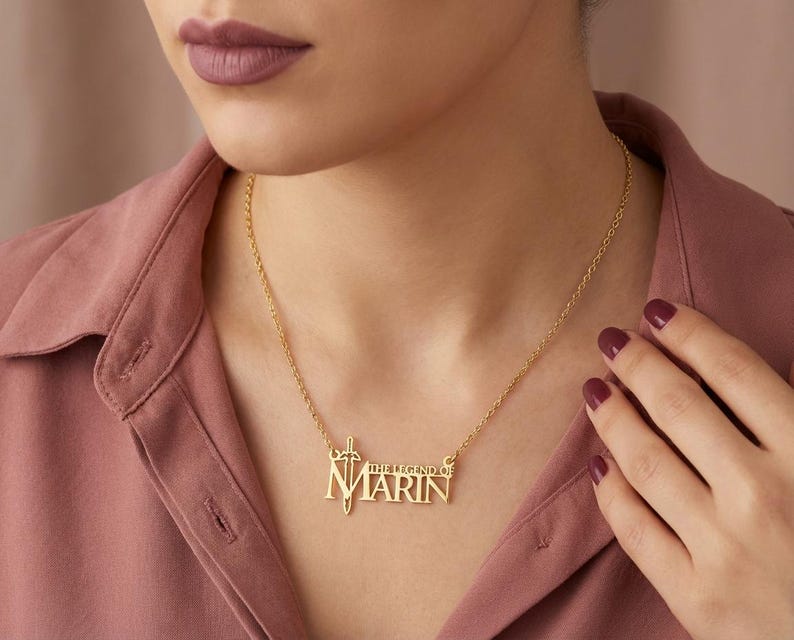 May include: A gold-colored necklace with the name "Marin" in block letters, with the words "THE LEGEND OF" above it. The necklace has a sword and shield design on the left side. The necklace is worn on a person wearing a pink shirt.