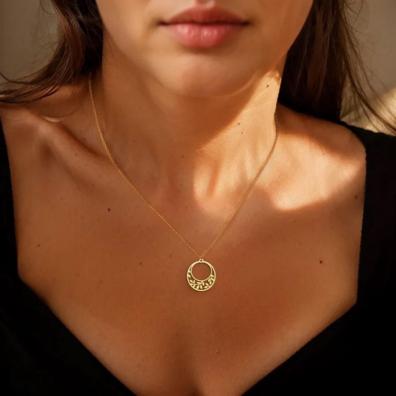 Puede incluir: Un collar dorado con un colgante circular con un dise&ntilde;o calado decorativo. El collar se lleva sobre un fondo oscuro, resaltando los intrincados detalles del colgante y la delicada estructura de la cadena. La joya es un accesorio sencillo pero elegante.