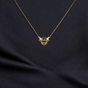 May include: A gold-colored necklace featuring a Highland cow pendant. The pendant has a stylized cow head design with horns and a name, "Raylen", inscribed in script. The necklace chain is delicate, and the pendant is centered.