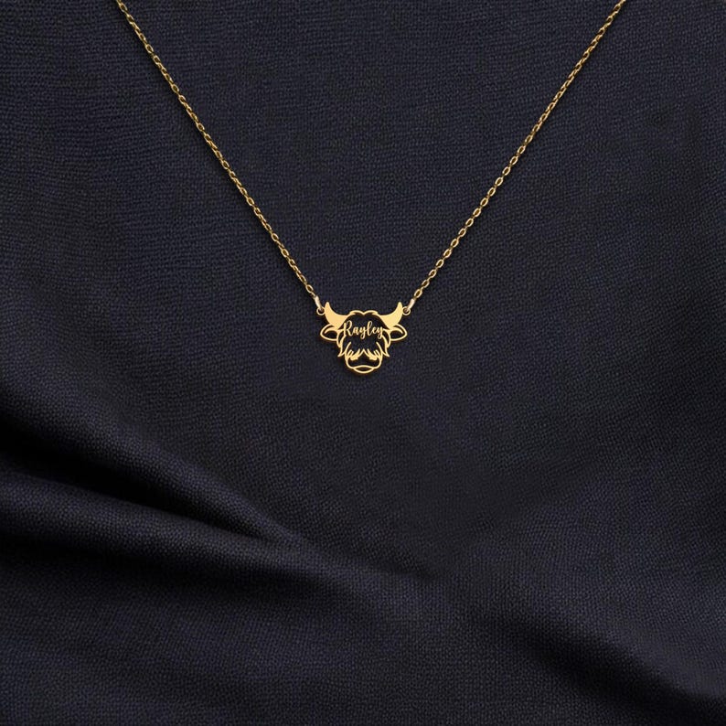 May include: A gold-colored necklace featuring a Highland cow pendant. The pendant has a stylized cow head design with horns and a name, "Raylen", inscribed in script. The necklace chain is delicate, and the pendant is centered.