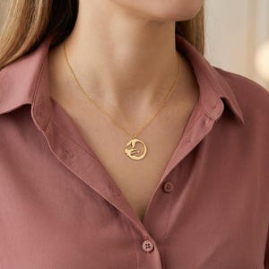 May include: A gold-colored pendant necklace featuring a circular design with a stylized figure. The necklace is worn on a person wearing a rose-colored button-down shirt. The pendant hangs from a delicate chain.