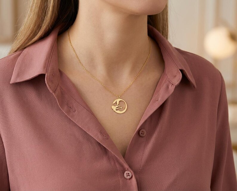 May include: A gold-colored pendant necklace featuring a circular design with a stylized figure. The necklace is worn on a person wearing a rose-colored button-down shirt. The pendant hangs from a delicate chain.