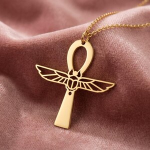 May include: A gold-colored Ankh pendant necklace. The pendant features a stylized cross with a loop at the top and detailed wings. The necklace is displayed on a pink velvet surface, highlighting the jewelry's design and color.