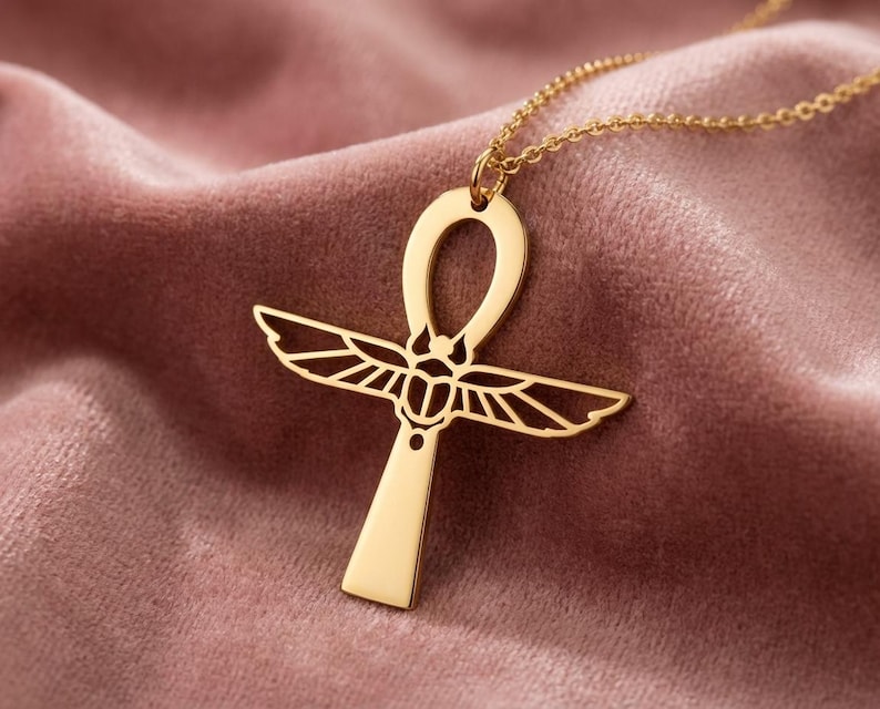 May include: A gold-colored Ankh pendant necklace. The pendant features a stylized cross with a loop at the top and detailed wings. The necklace is displayed on a pink velvet surface, highlighting the jewelry's design and color.