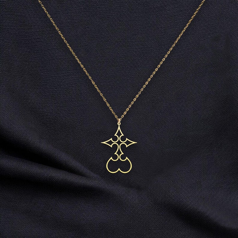 Puede incluir: Collar con colgante dorado con un dise&ntilde;o de cruz estilizada. El colgante cuelga de una delicada cadena sobre un fondo oscuro. La cruz tiene cortes sim&eacute;tricos y una base en forma de coraz&oacute;n.