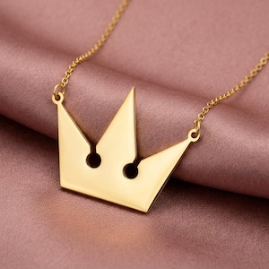 May include: A gold-colored crown pendant necklace. The pendant has a minimalist design with two circular cutouts and is suspended from a delicate chain. The necklace is displayed on a pink satin surface.