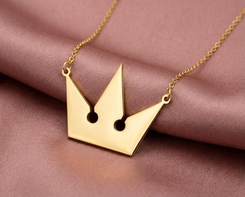 May include: A gold-colored crown pendant necklace. The pendant has a minimalist design with two circular cutouts and is suspended from a delicate chain. The necklace is displayed on a pink satin surface.