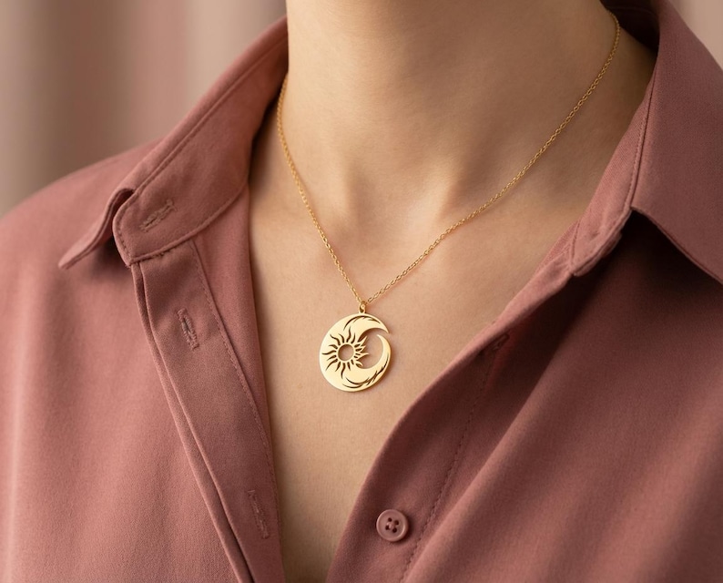 Op de afbeelding: Een goudkleurige hangerketting met een zon- en maandesign. De ronde hanger heeft een gedetailleerd zonnepatroon en een gebogen maanvorm. De ketting wordt gedragen over een roze overhemd.