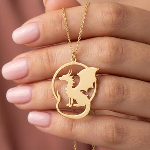 May include: A gold-colored necklace featuring a dragon pendant. The dragon is inside a circular frame, with its wings spread. The necklace is held up by a delicate chain. The dragon is a mythical creature with a long tail and wings.
