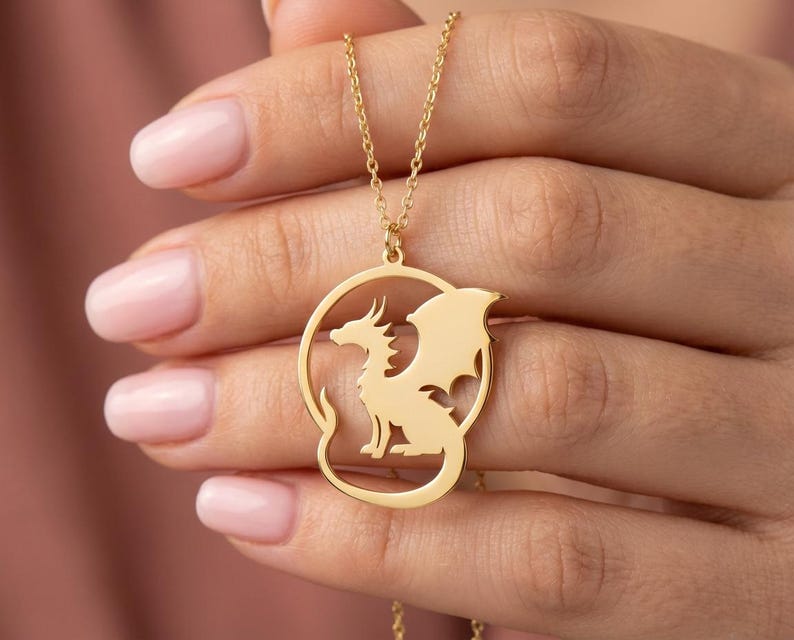 May include: A gold-colored necklace featuring a dragon pendant. The dragon is inside a circular frame, with its wings spread. The necklace is held up by a delicate chain. The dragon is a mythical creature with a long tail and wings.