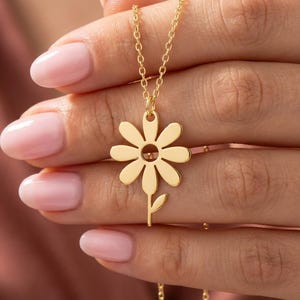 May include: A gold daisy pendant necklace. The pendant has a daisy flower design with a stem and leaf. The necklace is on a fine chain.