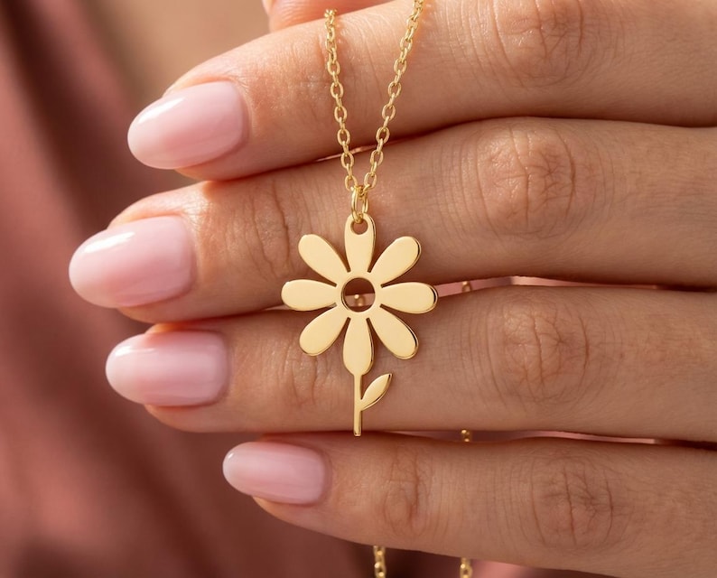 May include: A gold daisy pendant necklace. The pendant has a daisy flower design with a stem and leaf. The necklace is on a fine chain.