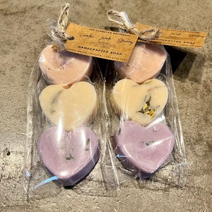 May include: Three heart-shaped bars of handcrafted soap wrapped in clear plastic. The soap is lavender, cream, and pink. The label reads "The Charlton Artisanal Crafted Love Story Handmade Soap Small Batch | Natural | Cruelty Free"