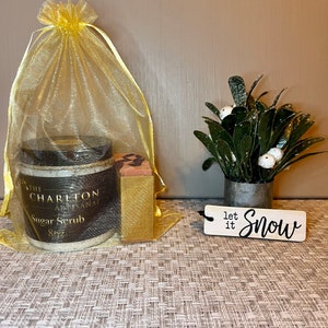 Puede incluir: Un set de regalo con un tarro de exfoliante de azúcar etiquetado como "The Charlton Artisan Sugar Scrub 8oz", una pequeña pastilla de jabón, una bolsa de organza amarilla y un letrero de madera que dice "Let it Snow".