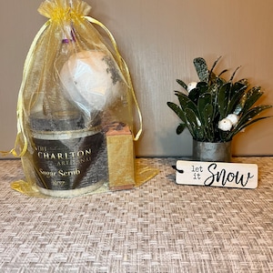 May include: A gift set in a gold organza bag containing a jar of sugar scrub, a bath bomb, and a bar of soap. The jar is labelled "The Charlton Artisanal Sugar Scrub 8oz". A small wooden sign reads "let it Snow".