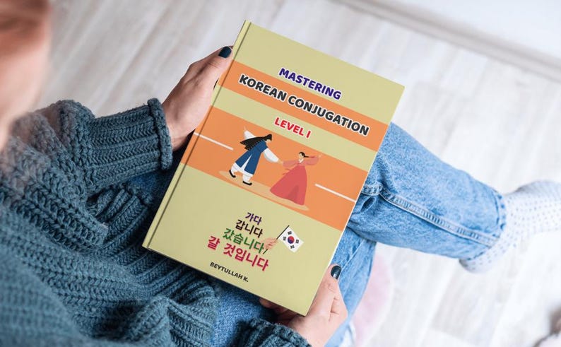 May include: A book titled Mastering Korean Conjugation Level 1 with a light yellow cover featuring an illustration of two people in traditional clothing, a small South Korean flag, and Korean text including Gada, Gapnida, Gatseumnida, and Gal Geosimnida.