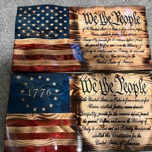 May include: Two rectangular wooden wall plaques featuring a wavy, scorched finish. The top plaque displays a blue field with white stars and the Preamble to the U.S. Constitution. The bottom plaque shows a blue field with 13 stars, 1776, and the Preamble.