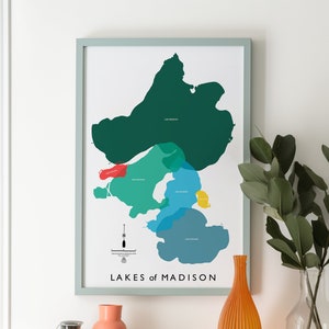 Puede incluir: Una impresión enmarcada de un mapa de los Lagos de Madison, Wisconsin. El mapa está en un marco azul claro y presenta una ilustración colorida de los lagos. La impresión está sobre una mesa de madera con un jarrón de vegetación y otros artículos decorativos.