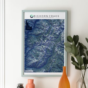 May include: A framed map of the Bighorn Crags in Idaho, with a blue and green color scheme. The map is hanging on a white wall above a wooden table with a vase, a jar, and a stack of books.