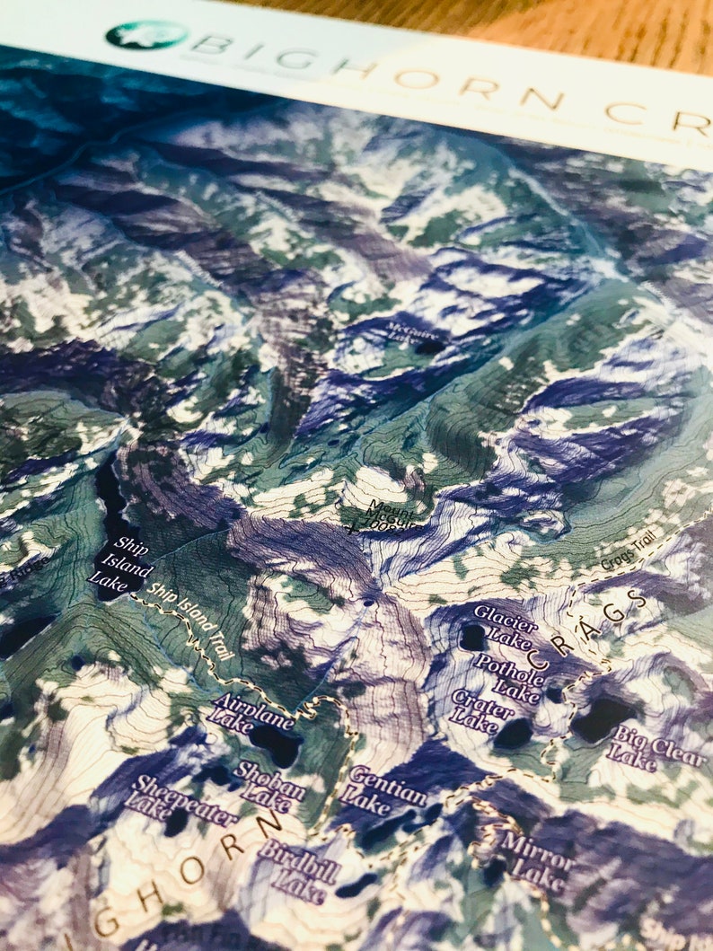 May include: A detailed map of the Bighorn Crags area in Wyoming, showing various trails, lakes, and mountains. The map features a blue and green color scheme with contour lines indicating elevation changes.