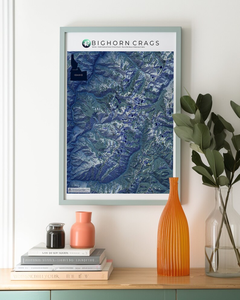 May include: A framed map of the Bighorn Crags in Idaho, with a blue and green color scheme. The map is hanging on a white wall above a wooden table with a vase, a jar, and a stack of books.