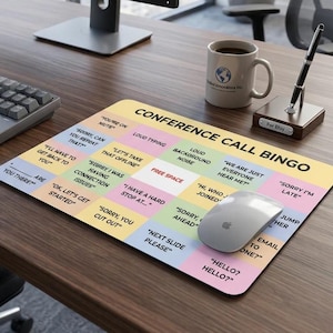 May include: A rectangular desk mat featuring a Conference Call Bingo game grid with pastel-coloured squares. Each square contains common meeting phrases like You're on mute, Loud typing, and Free space. The mat is placed on a wooden desk surface.