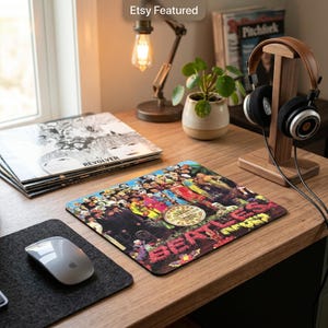 May include: A square mouse pad featuring the colourful Sgt. Pepper's Lonely Hearts Club Band album cover art by The Beatles. The design displays a collage of various figures and the band name in red floral lettering against a dark background.