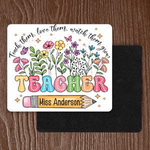 May include: White background with colorful flowers and the word "TEACHER" in rainbow letters. A yellow pencil with a pink eraser is below the word "TEACHER" with the text "Miss Anderson" on it. The text "Teach them, love them, watch them grow!" is above the flowers.