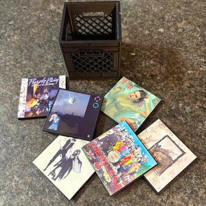 May include: A black plastic crate with several square coasters featuring album art. The coasters showcase album covers with vibrant colours and various designs, including text and images. The coasters are arranged on a dark surface.
