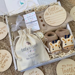 Pode incluir: Uma caixa de presente cheia de itens para beb&ecirc;s, incluindo um macac&atilde;o bege, um par de botinhas marrons com carinhas de girafa, uma pequena bolsa de cord&atilde;o branco com o texto "Little & Loved Keepsake Baby Milestones", e v&aacute;rios c&iacute;rculos de madeira com frases como "I can Roll Over", "I can Smile", "10+ Months", "My First Tooth", e "Little & Loved".
