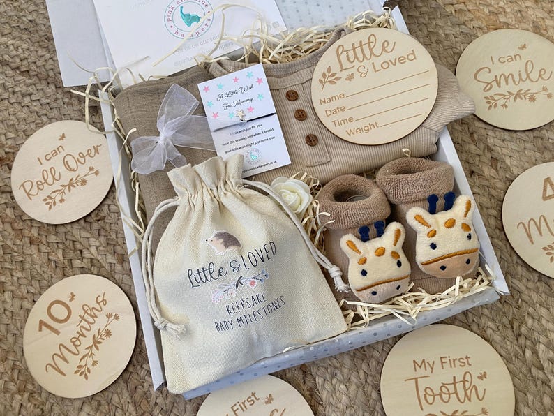 Pode incluir: Uma caixa de presente cheia de itens para beb&ecirc;s, incluindo um macac&atilde;o bege, um par de botinhas marrons com carinhas de girafa, uma pequena bolsa de cord&atilde;o branco com o texto "Little & Loved Keepsake Baby Milestones", e v&aacute;rios c&iacute;rculos de madeira com frases como "I can Roll Over", "I can Smile", "10+ Months", "My First Tooth", e "Little & Loved".