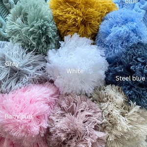 May include: A collection of knitted hats featuring large yarn pom-poms in various colors including sage green, mustard, baby blue, grey, white, steel blue, baby pink, dusky pink, and biscuit, with text labels identifying each color shade.